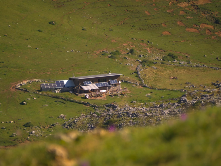 Aerial view of a sustainable cabin with solar panels, nestled in a vast, green hillside landscape.