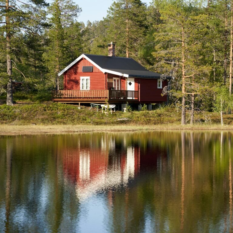 Charming red cabin reflected in a serene lake in Krøderen, Norway, amidst lush greenery.